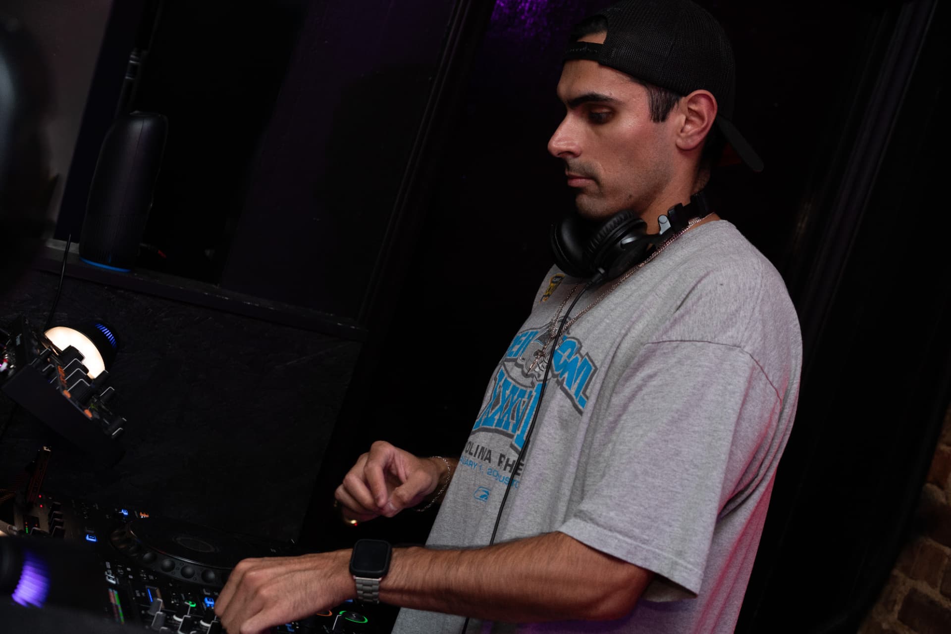 BINO behind the decks, headphones around his neck, dark club lighting