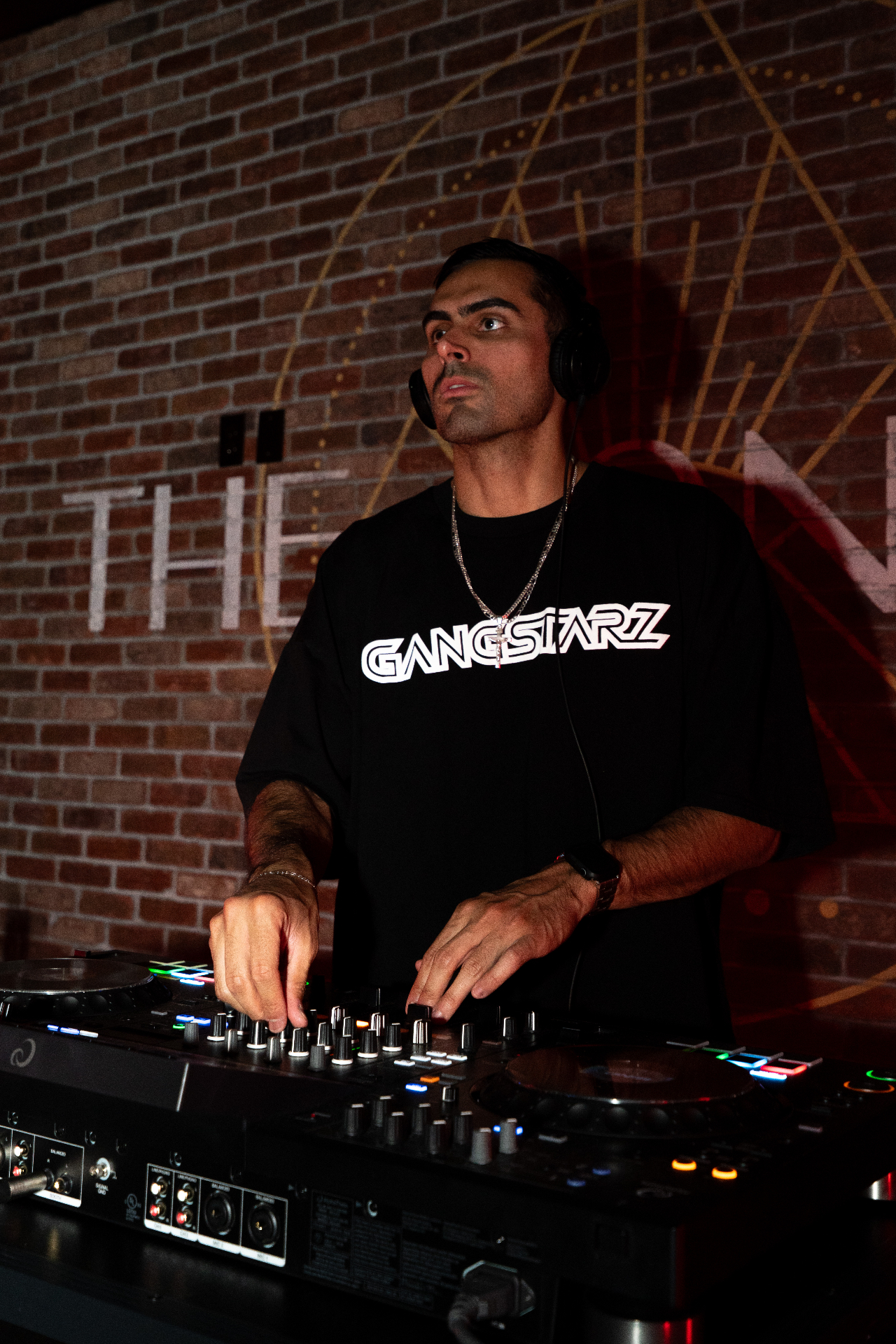 BINO behind the decks in a Gangstarz tee, headphones on, brick-wall backdrop